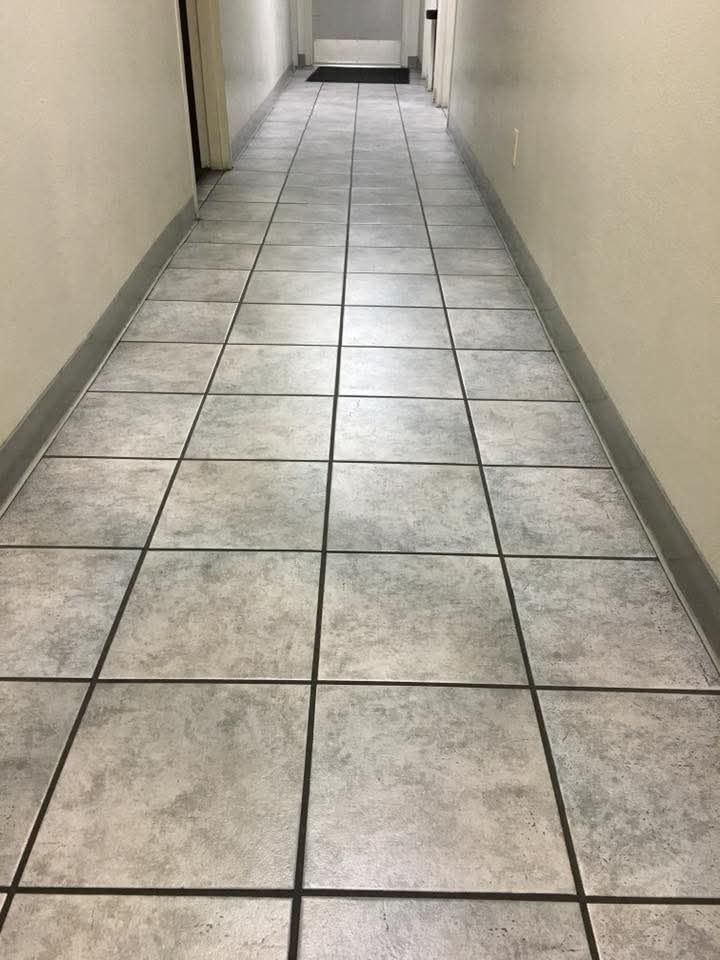 Long, narrow hallway with grey square floor tiles and plain walls leading to a door.